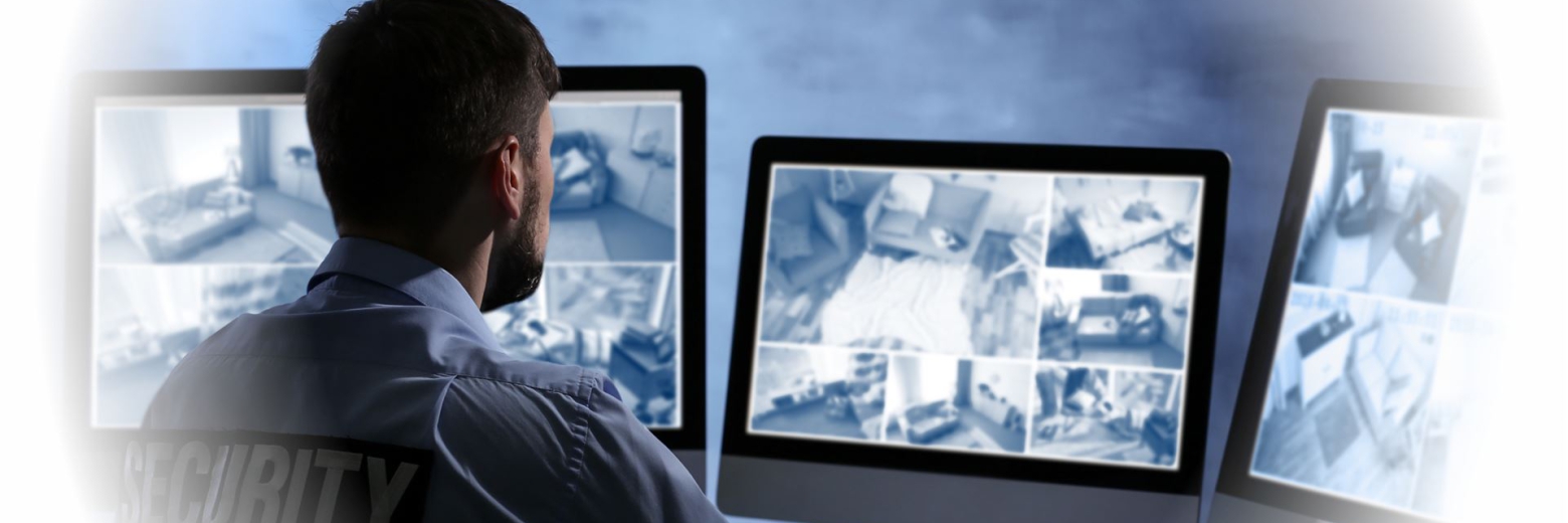 Ein Mann überwacht von einem Schreibtisch aus multiple Computermonitore, auf denen Überwachungskamera-Feeds laufen.