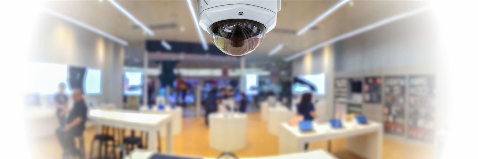 Eine Überwachungskamera zeigt den weiten Innenraum eines belebten, modernen Einzelhandelsgeschäfts mit Kleiderständern und Kunden.